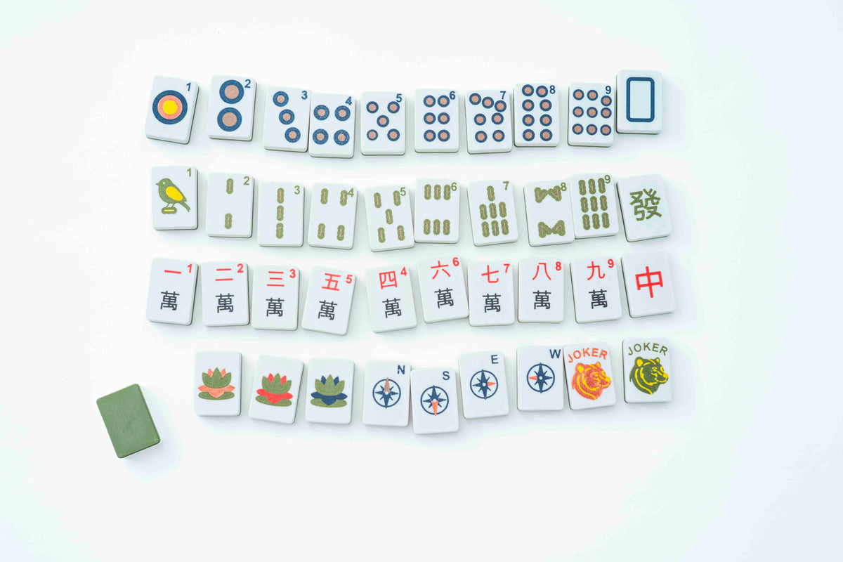 A set of Mahjong tiles arranged in rows on a plain white background, with a small green accessory piece placed near the lower left.
