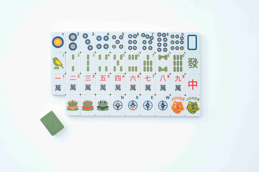 A set of Mahjong tiles arranged in a grid on a white background, with a single green tile placed to the lower left. The tiles feature traditional Chinese characters, numbers, and colorful illustrations including birds, lotus flowers, compasses, and jokers.
