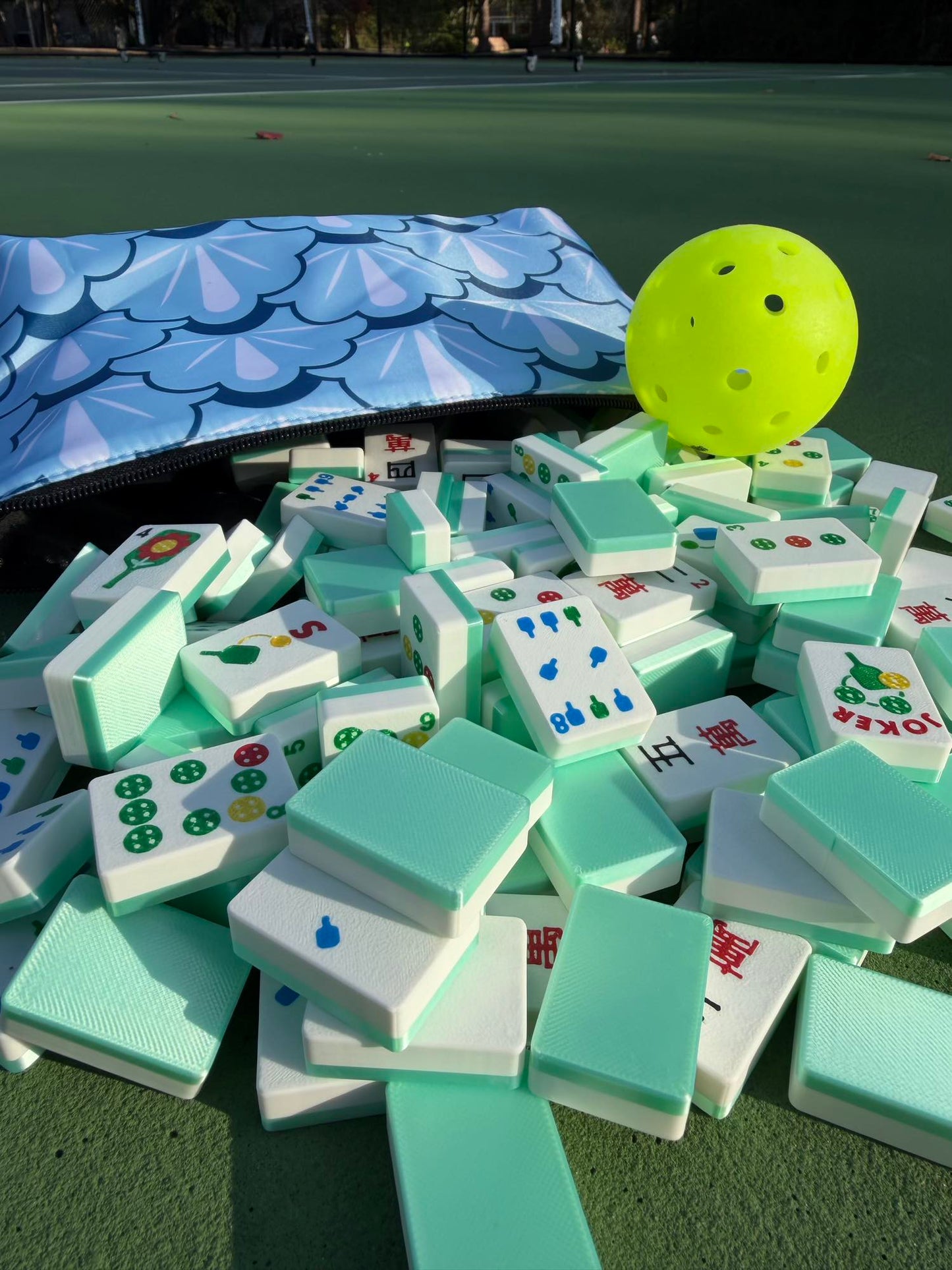 A yellow pickleball rests atop a mint-green pile of Travel Mahjong Tiles spilling from a scalloped pouch on a tennis court.
