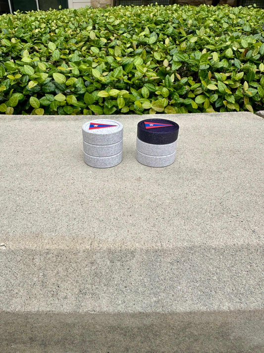 Two stacked trios of custom backgammon checkers sit on a ledge before a hedge, one topped white, one dark purple.