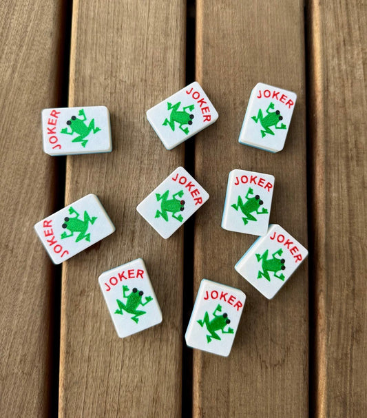Eight Custom Jokers - Frogs tiles, each stamped with a green frog, scatter across a wood deck.