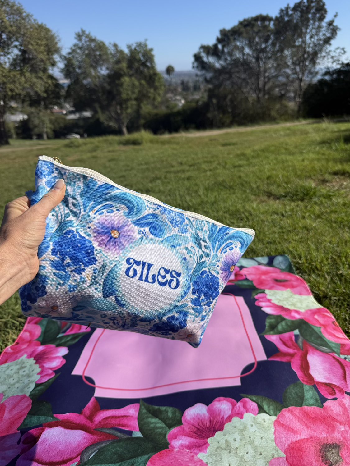 A hand holds a blue and white floral zippered pouch with the name 'TILES' printed in blue script on the front. The pouch is shown outdoors over a pink and green floral blanket on a grassy lawn with trees in the background.