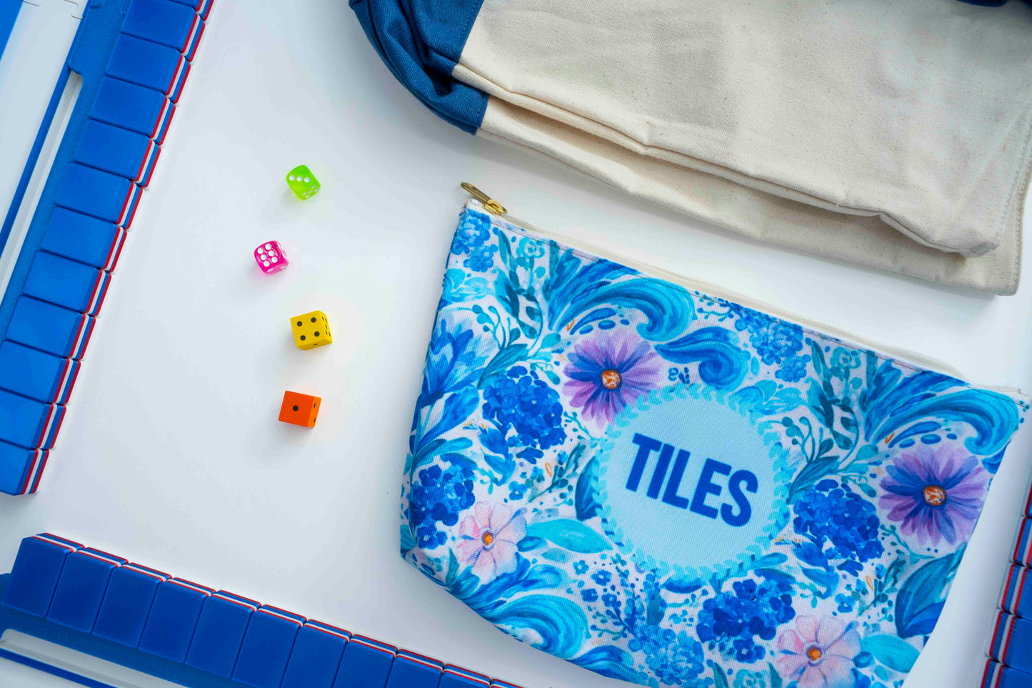 A personalized zippered pouch with a blue and white floral tile pattern, featuring the word 'TILES' in blue on a light blue circle. The pouch is styled in a flat-lay composition alongside a beige canvas bag, colorful dice, and blue game boards on a white surface.