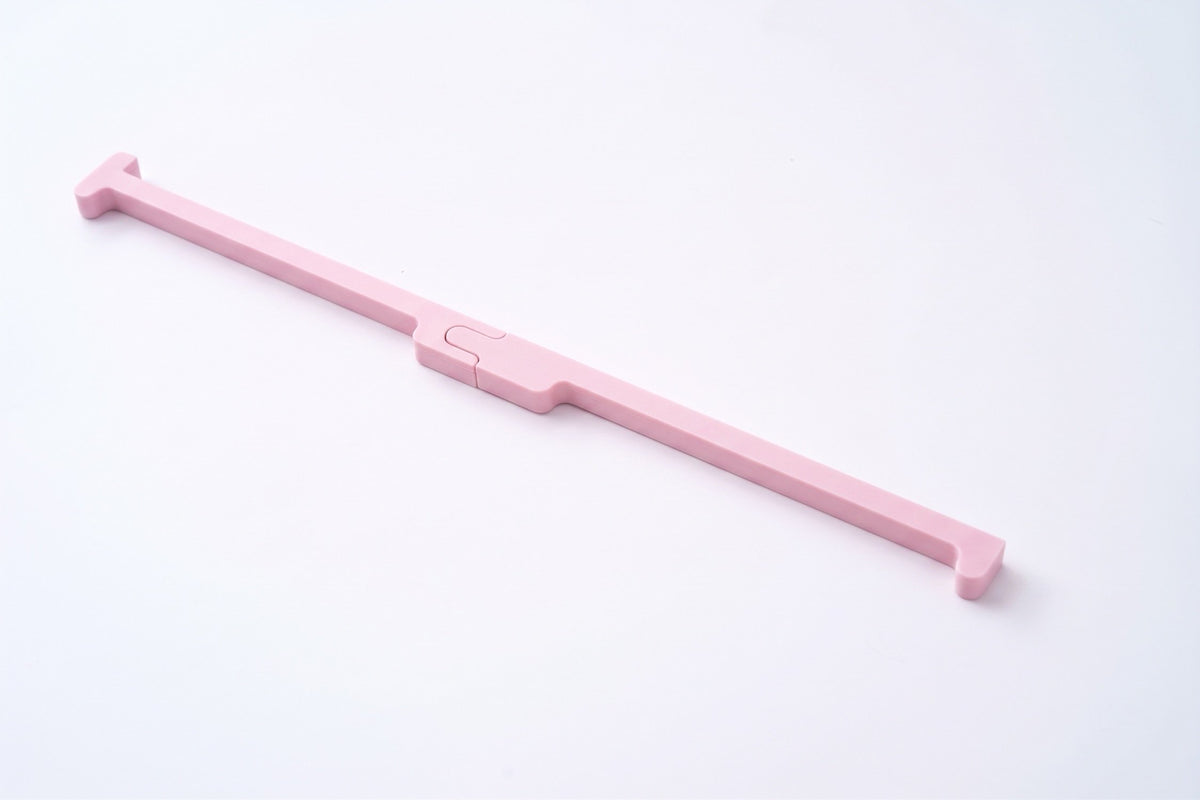 A light pink plastic tool with a long handle and a small central slider, photographed diagonally on a clean white background.