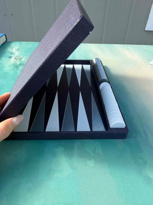 A hand holding open a dark gray foam presentation box containing a black and white backgammon board and a white-and-black cylindrical game piece, set on a mint green tabletop.