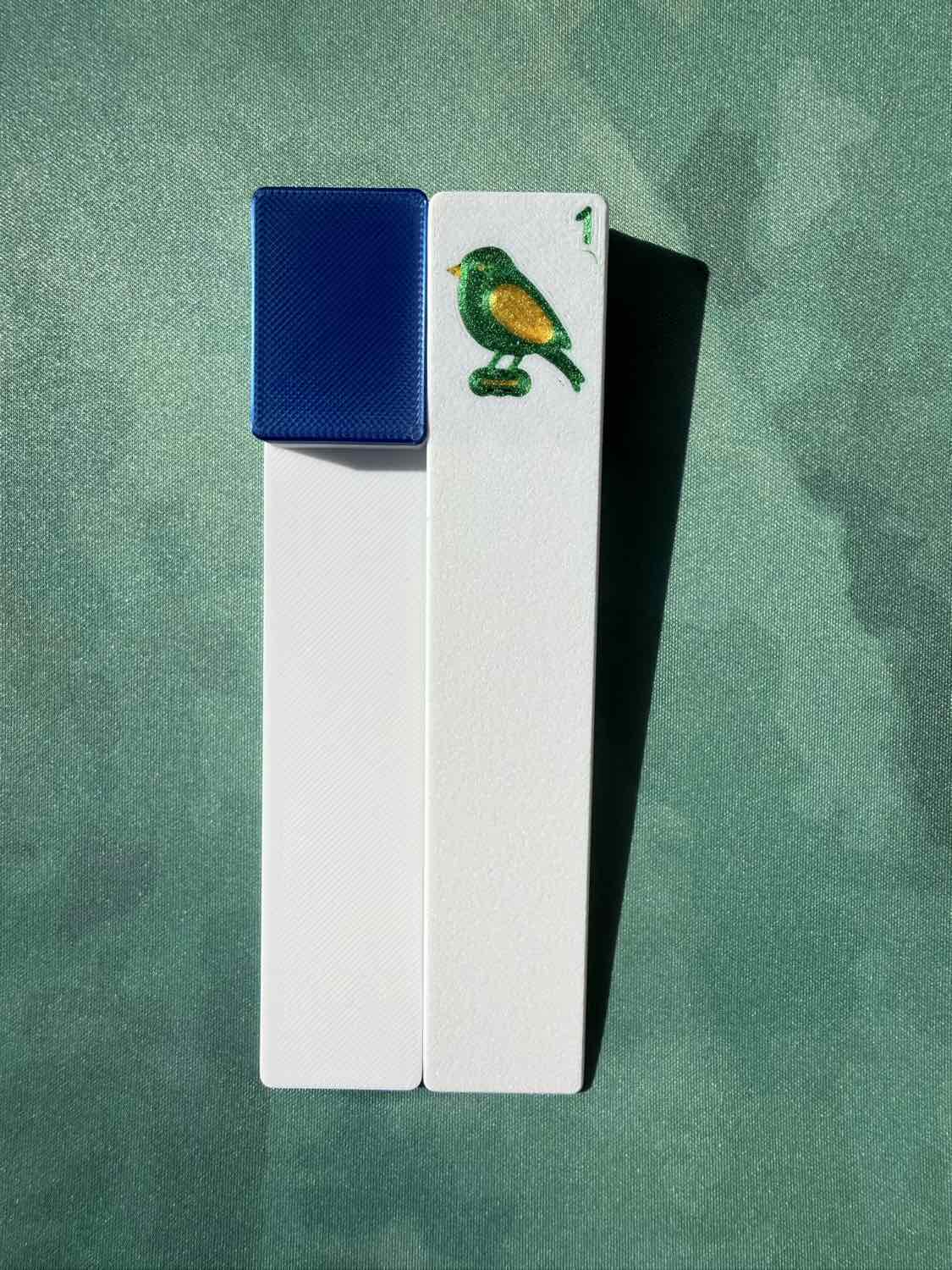 Two white rectangular cards or tiles standing upright on a textured green background. One card has a textured blue square on the left side, and the other has a small painted bird in green, yellow, and blue near the top right.