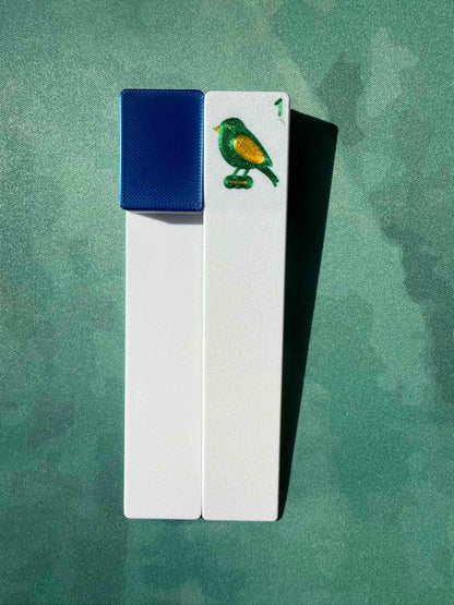 Two white rectangular cards or tiles standing upright on a textured green background. One card has a textured blue square on the left side, and the other has a small painted bird in green, yellow, and blue near the top right.
