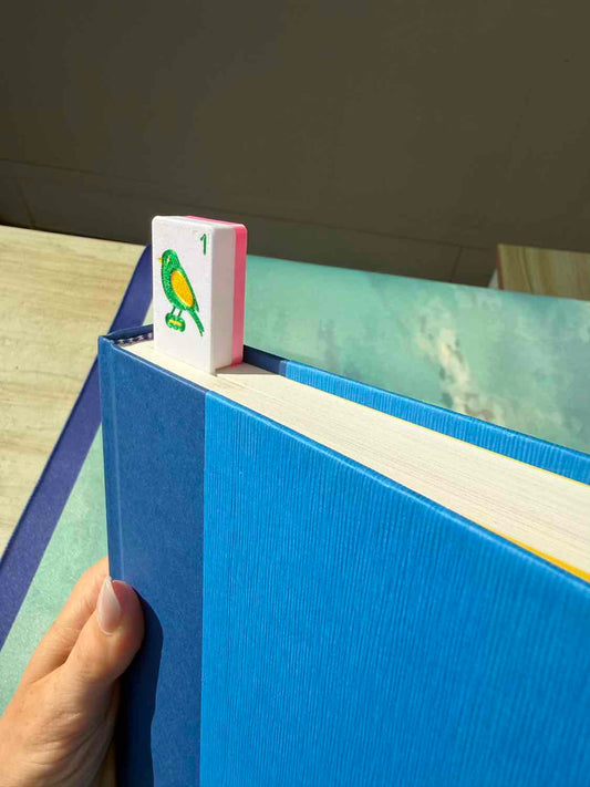 A close-up of a blue hardcover book held by a hand, with a small white bookmark featuring a green and yellow bird and the number 1 tucked between the pages.