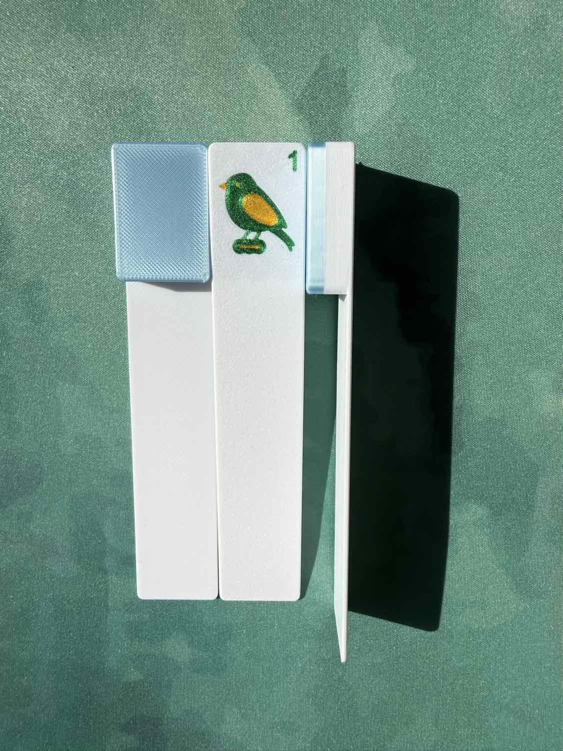 A white rectangular card or tool with a small bird illustration and the number 1, shown alongside a black rectangular piece on a textured green background.