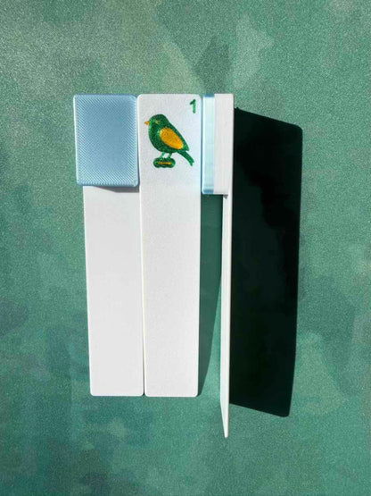 A white rectangular card or tool with a small bird illustration and the number 1, shown alongside a black rectangular piece on a textured green background.