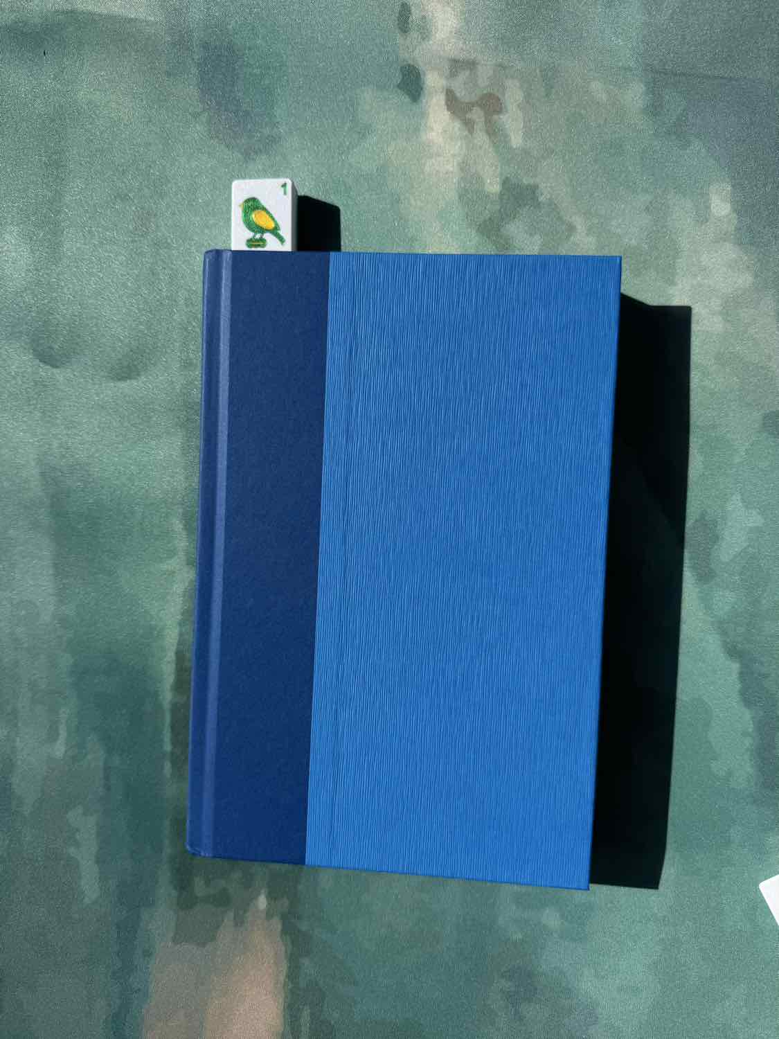A hardcover notebook or journal with a textured blue cover, featuring a solid dark blue spine band and a small white bookmark card with a green bird illustration, placed on a mottled green surface.