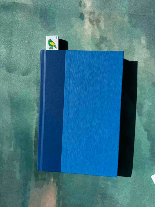 A hardcover notebook or journal with a textured blue cover, featuring a solid dark blue spine band and a small white bookmark card with a green bird illustration, placed on a mottled green surface.