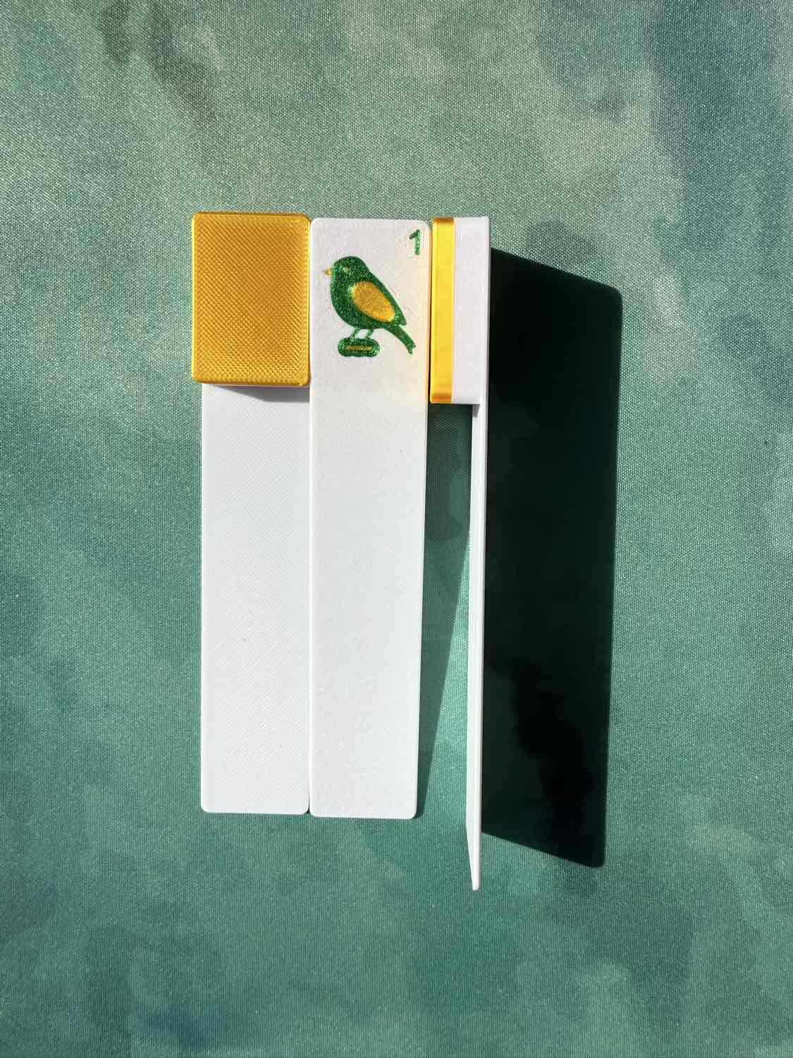 A set of three playing cards (one face-up showing a green and yellow bird with a '1' and a gold border, two cards face down) arranged on a textured green background with a strong shadow.