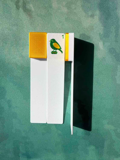 A set of three playing cards (one face-up showing a green and yellow bird with a '1' and a gold border, two cards face down) arranged on a textured green background with a strong shadow.