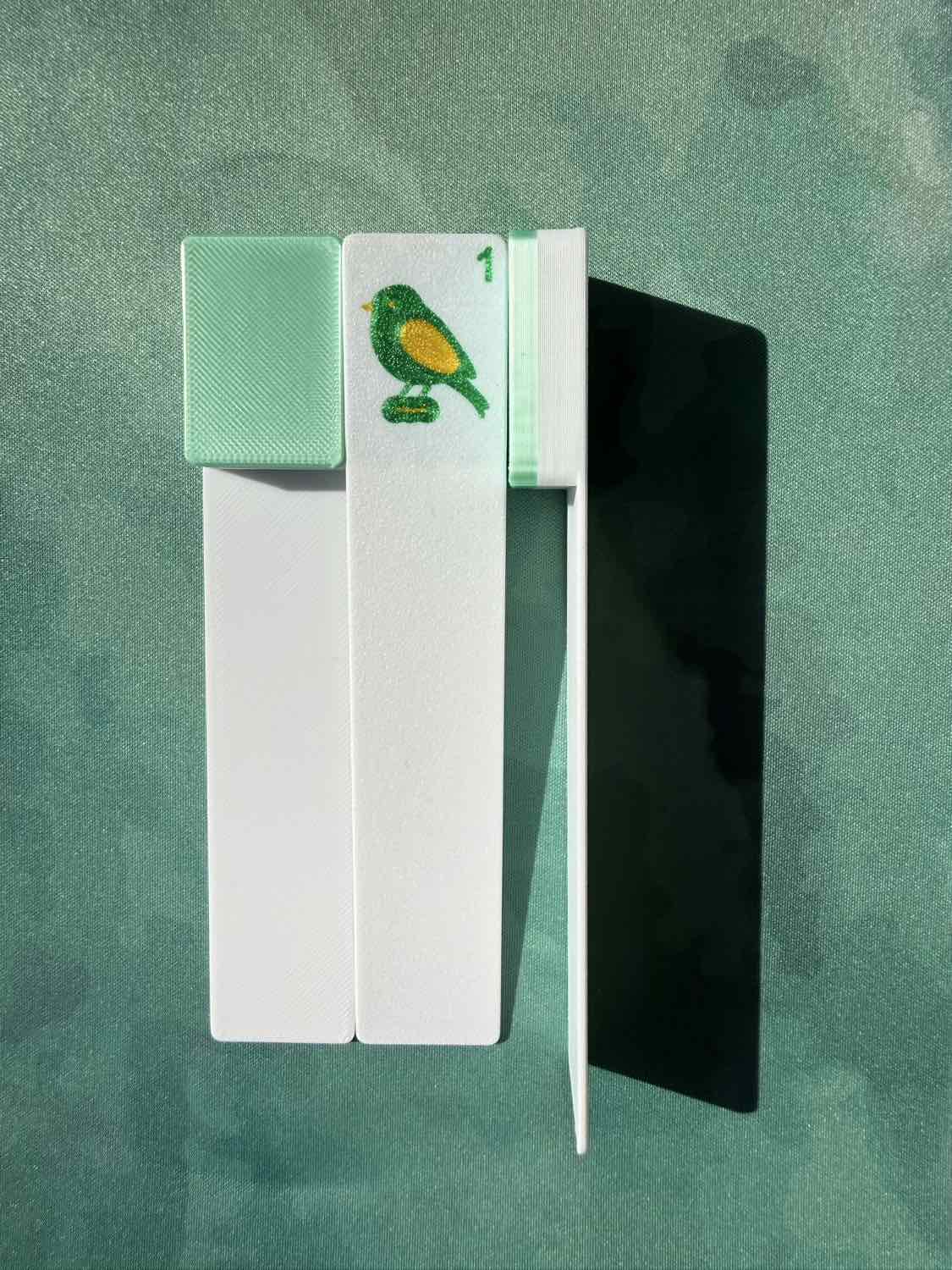 A set of mahjong tiles shown on a textured green background, featuring white backs, a mint green tile with a green and yellow bird, a black tile, and a white tile with a green and yellow bird.