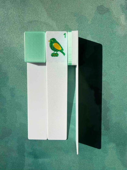 A set of mahjong tiles shown on a textured green background, featuring white backs, a mint green tile with a green and yellow bird, a black tile, and a white tile with a green and yellow bird.