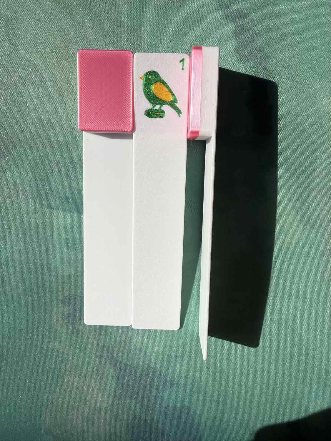 A white rectangular mahjong tile bookmark with a pink textured cap and a small bird illustration, placed on a green textured surface with a strong shadow.