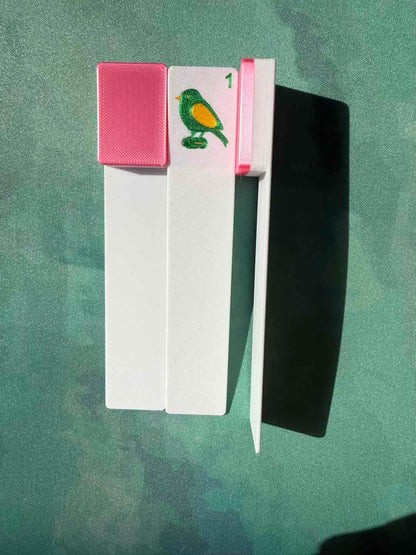 A white rectangular mahjong tile bookmark with a pink textured cap and a small bird illustration, placed on a green textured surface with a strong shadow.