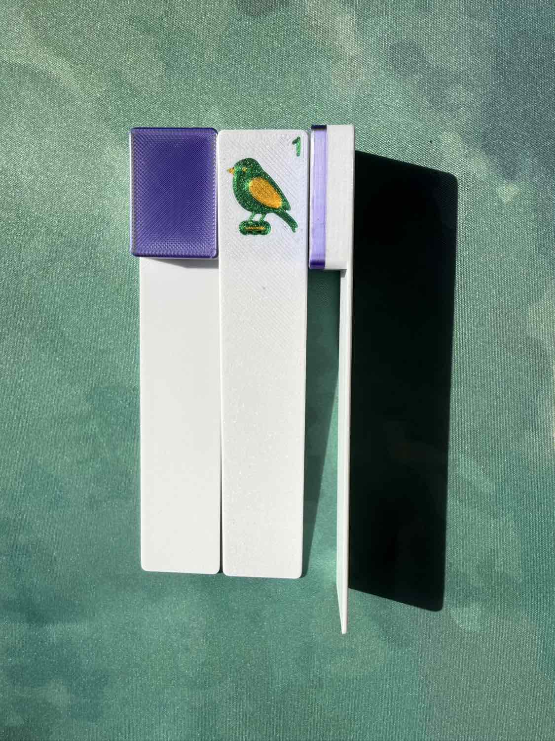 A pair of white wooden matchsticks with purple caps and a green bird emblem, displayed on a textured green background.