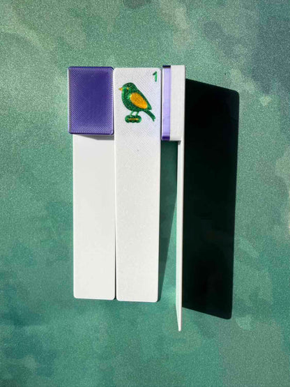 A pair of white wooden matchsticks with purple caps and a green bird emblem, displayed on a textured green background.
