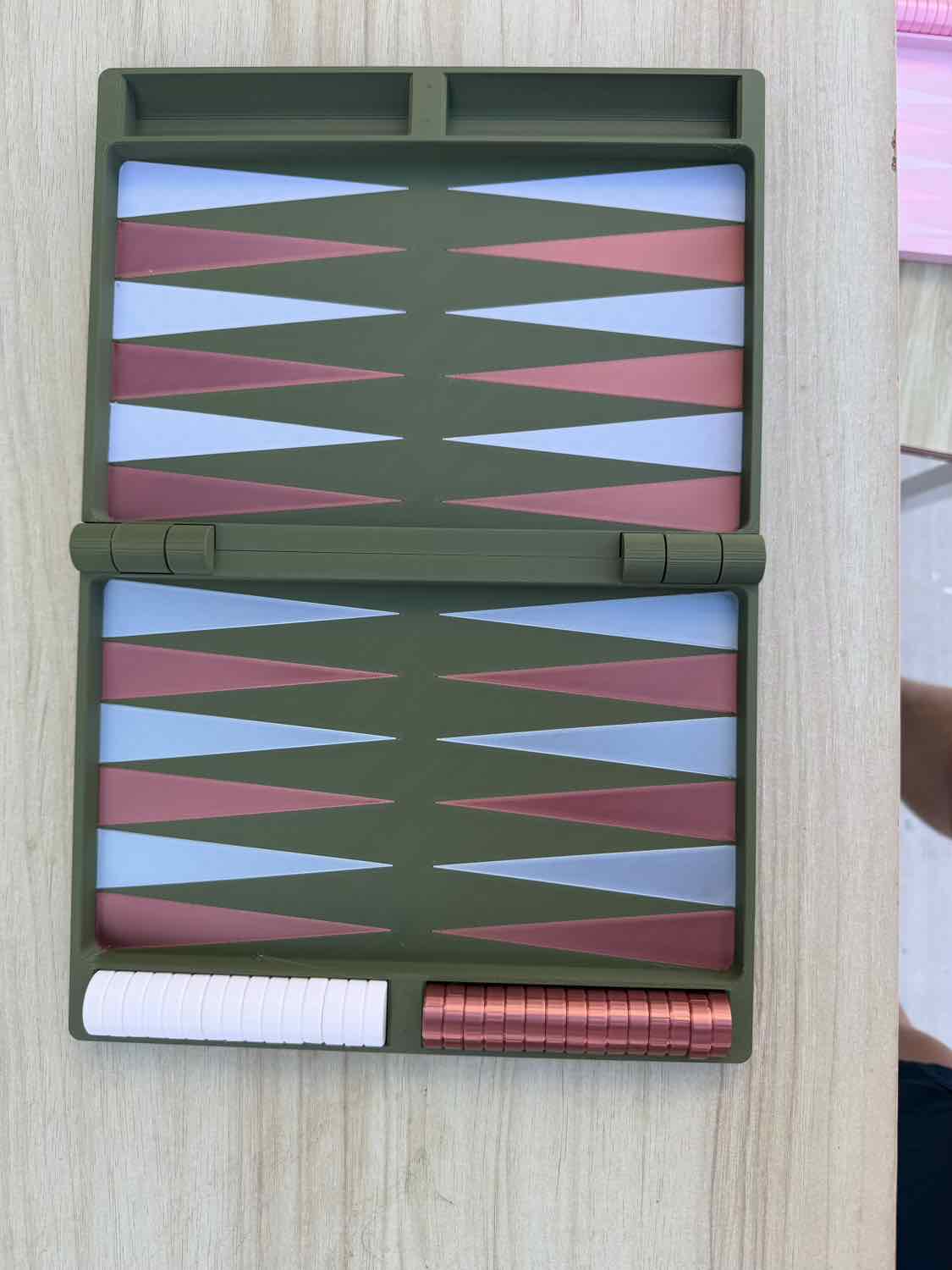 A compact folding backgammon board set shown open on a light wood surface. The board frame is olive green, with alternating pink and light blue triangles on both sides, plus a white pawn track and a row of pink game pieces.