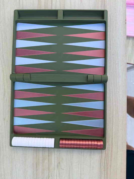 A compact folding backgammon board set shown open on a light wood surface. The board frame is olive green, with alternating pink and light blue triangles on both sides, plus a white pawn track and a row of pink game pieces.