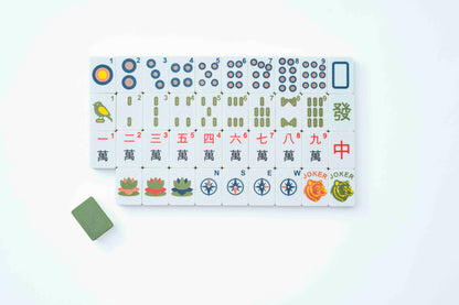 A set of Mahjong tiles arranged in a grid on a white background, with a single green tile placed to the lower left. The tiles feature traditional Chinese characters, numbers, and colorful illustrations including birds, lotus flowers, compasses, and jokers.