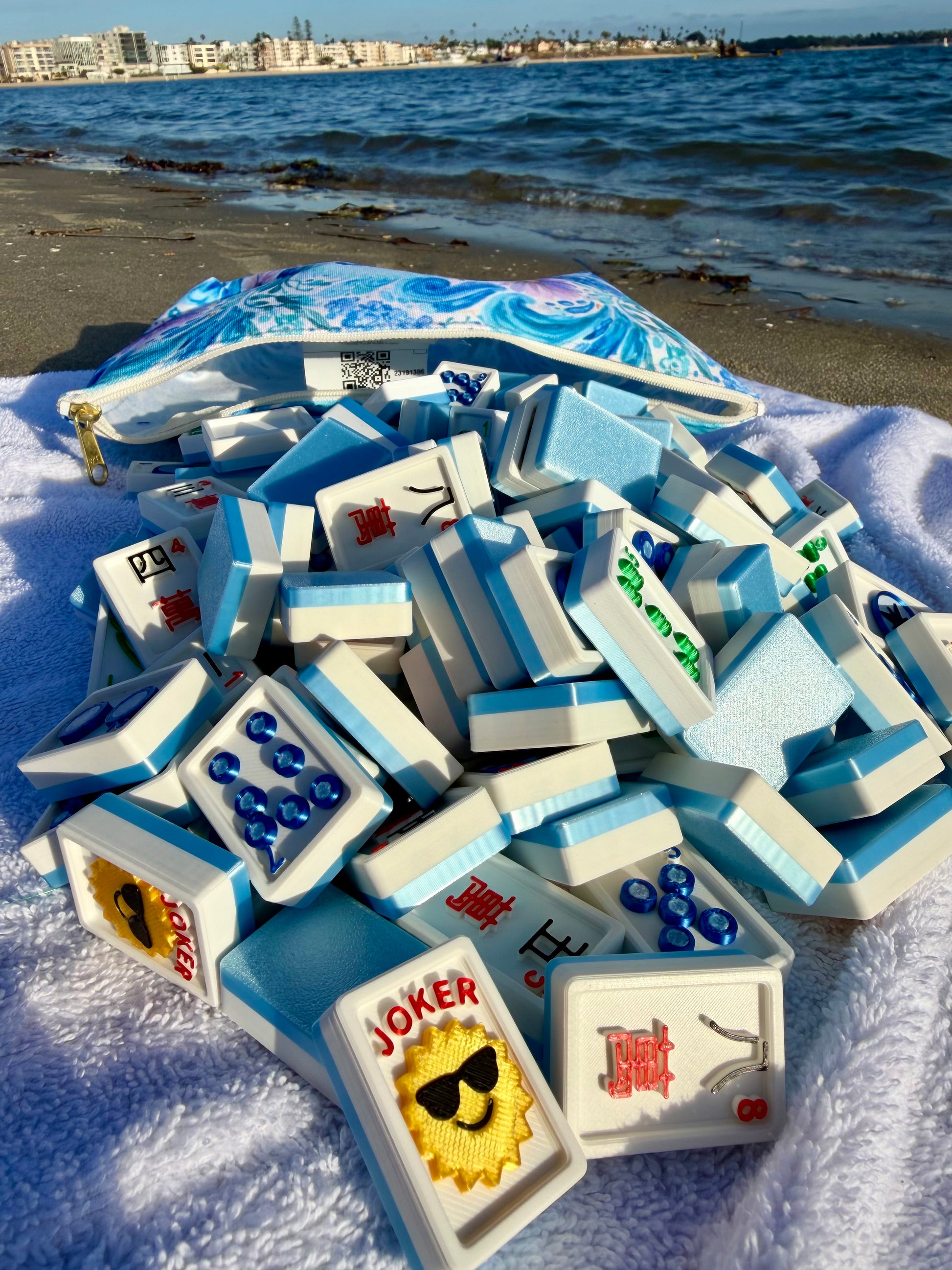 Light-blue Travel Mahjong Tiles - 3D spill from their pouch onto a white towel, a coastal skyline across the water.