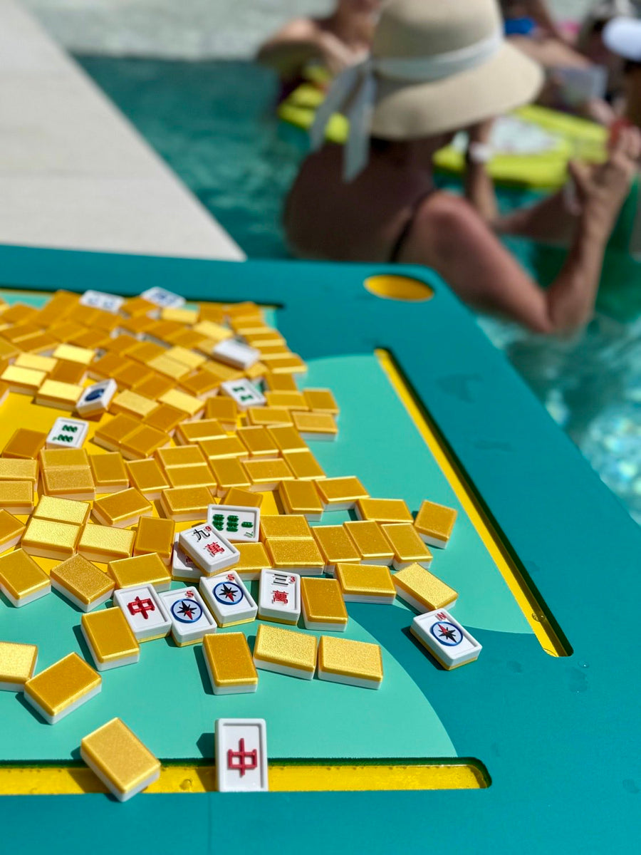 A close-up of a teal and yellow mahjong table with many loose yellow tiles and a few visible white tiles with Chinese characters, set by a swimming pool with people blurred in the background.