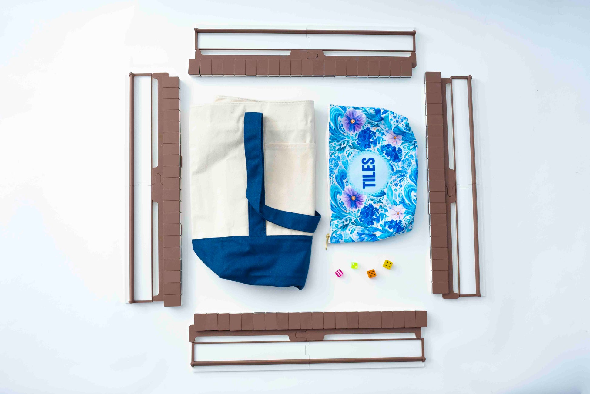 A flat-lay view of a DIY toy building set, featuring brown plastic wall panels, a cream canvas tote bag with blue straps, a blue and purple floral zippered pouch with the word 'TILES' printed on it, and a few small colorful plastic pieces, all arranged on a white background.