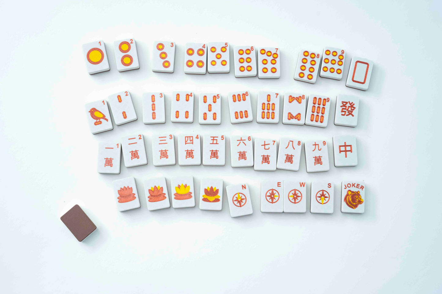 A set of Mahjong tiles arranged in rows on a clean white background, with a small brown rectangular accessory at the lower left. The tiles are white with orange and red markings and symbols.