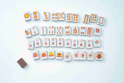 A set of Mahjong tiles arranged in rows on a clean white background, with a small brown rectangular accessory at the lower left. The tiles are white with orange and red markings and symbols.