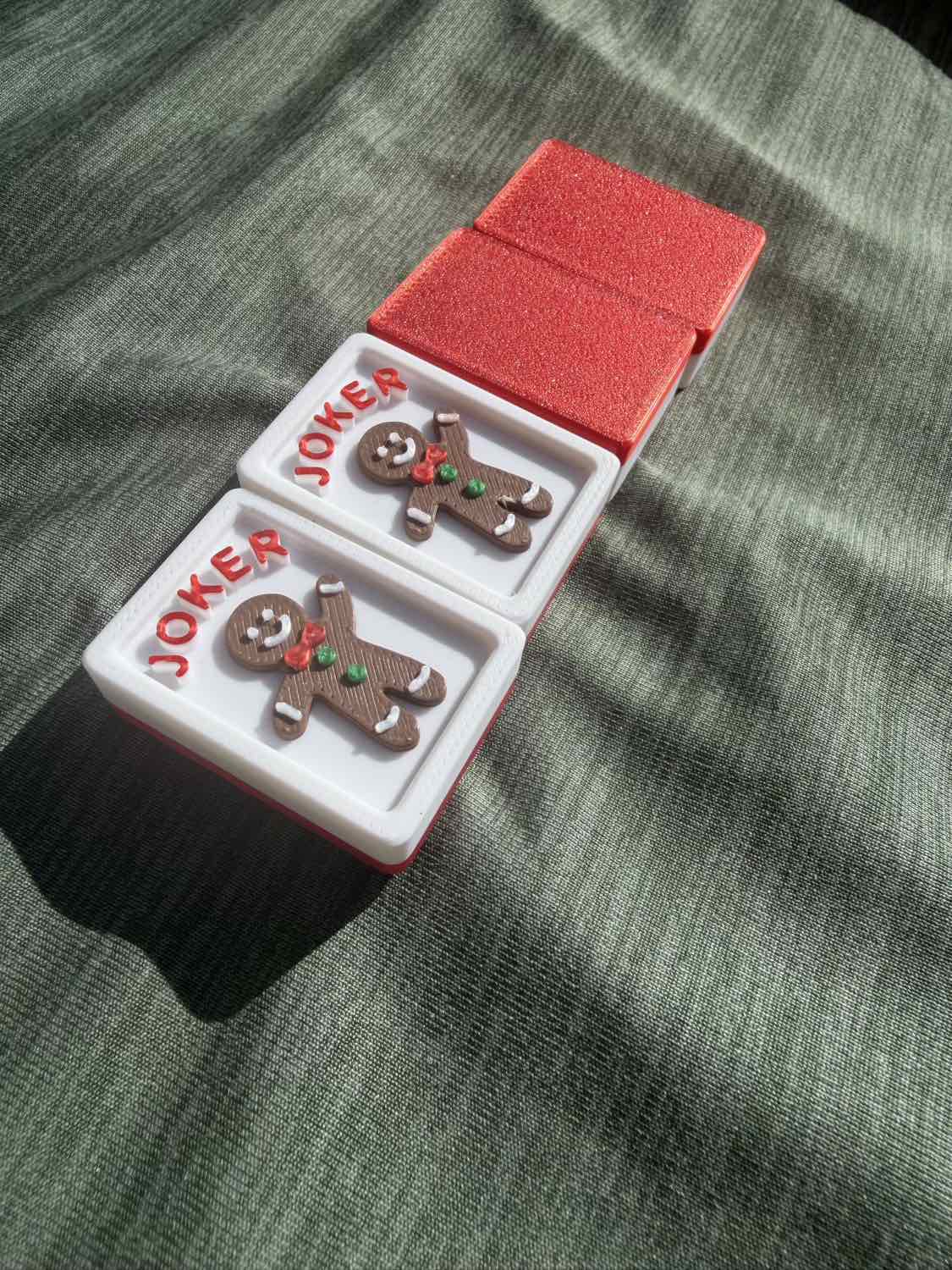 Two small Joker-themed gingerbread man Mahjong Tiles in white trays with red borders, placed on a red glittery box over a textured gray-green fabric background.