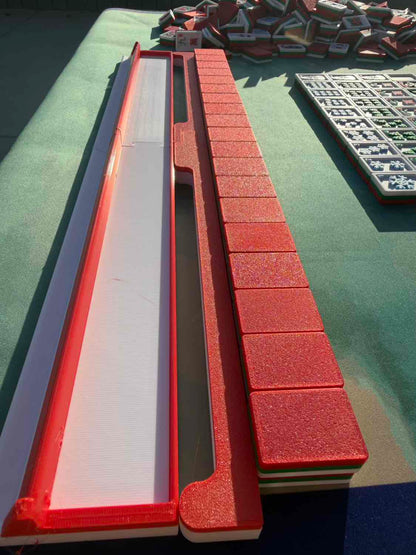 A close-up of a mahjong set showing a long red and white tile holder or tray alongside a row of red tile backs, with additional tiles and a rack visible in the background on a green surface.