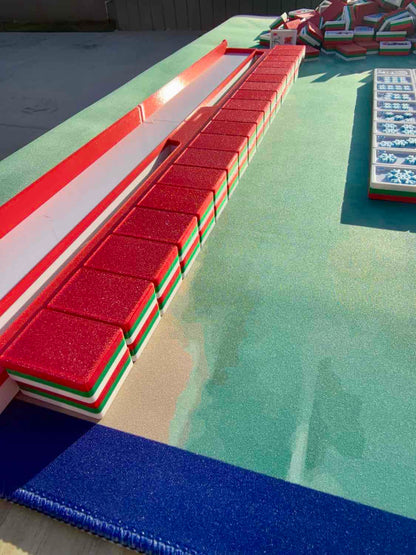 A set of Mahjong tiles arranged in a long row on a green felt playing surface, with a blue textured mat in the foreground and additional tiles visible on the right.