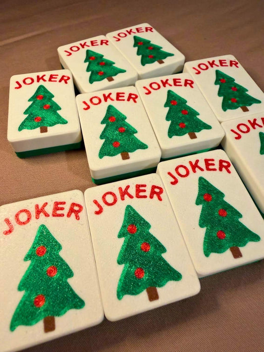 A set of white rectangular cards or tiles featuring a green Christmas tree with red ornaments and the word 'JOKER' in red, arranged on a brown fabric surface.
