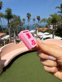 A hand holds a pink Flamingo crack tile with red characters, beach and palm trees behind