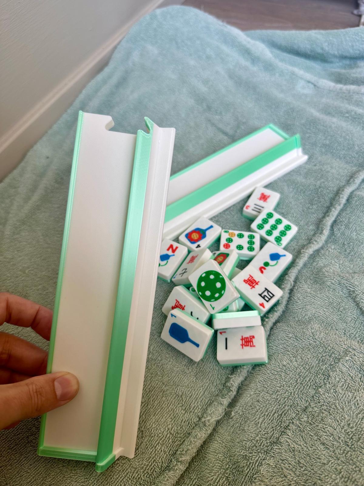 Hand holding a green and white floating mahjong rack beside loose pickleball-themed mahjong tiles on a towel surface.