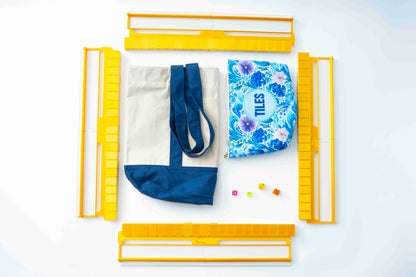 A flat-lay product presentation showing a canvas tote bag with a navy blue bottom and handles, a blue floral zippered pouch with the word 'TILES' printed on it, and several small colorful blocks, all arranged around a yellow folding frame on a white background.
