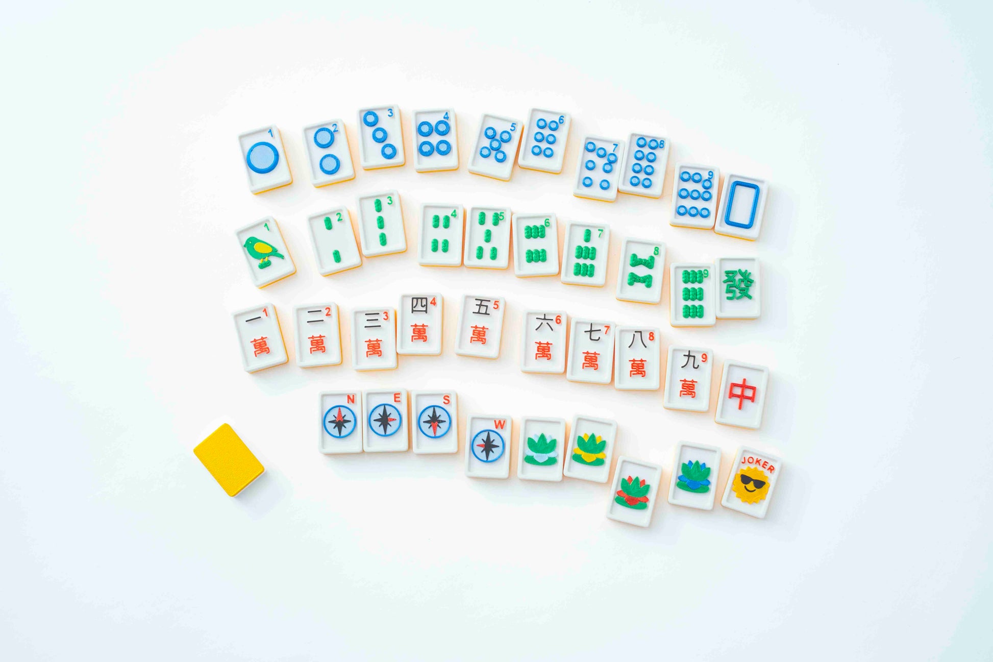 A set of Mahjong tiles arranged on a clean white background, with a small yellow eraser placed to the left. The tiles are mostly white with colorful printed symbols and characters in blue, green, red, and yellow.