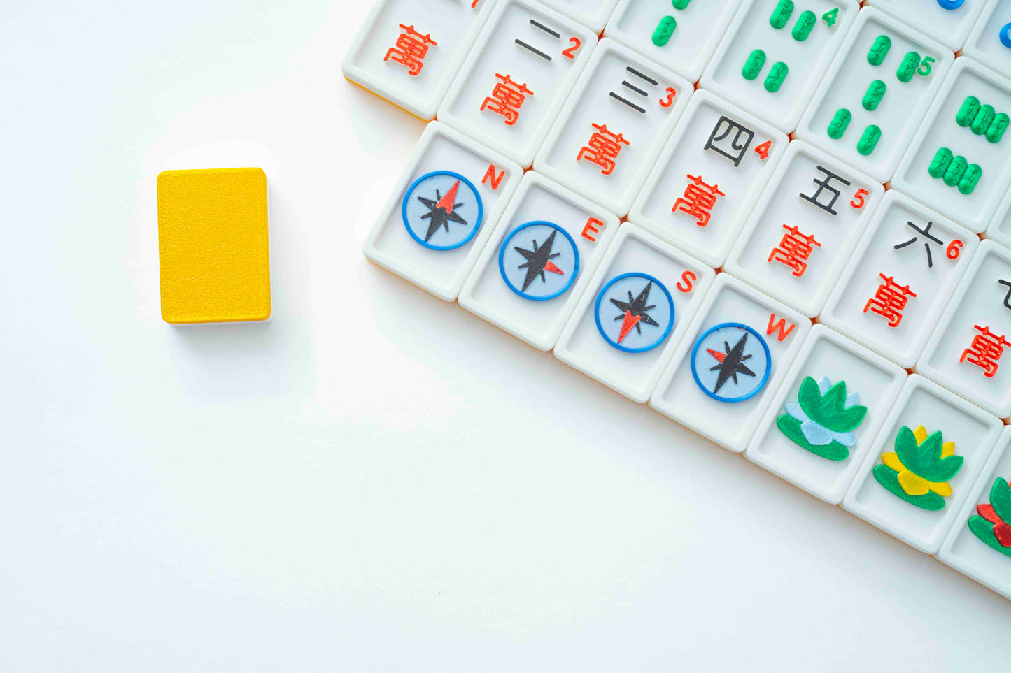 A close-up of Mahjong tiles arranged on a white surface, with a single yellow tile standing upright to the left. The visible tiles are white with red Chinese characters, green and yellow symbols, and blue compass markings.