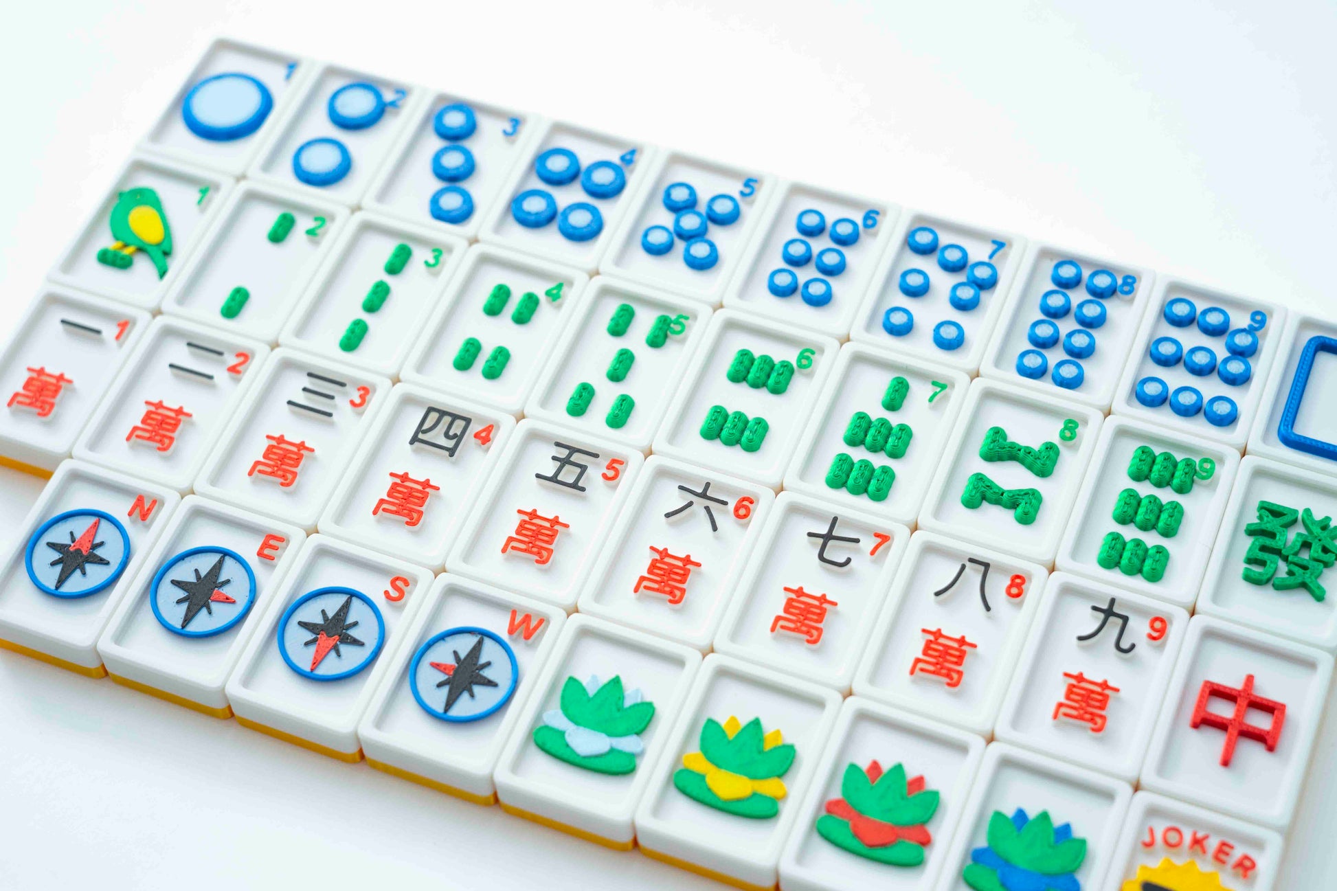 A close-up, angled view of a Mahjong game board showing multiple white tiles with colorful Chinese characters and symbols, photographed on a clean white background.