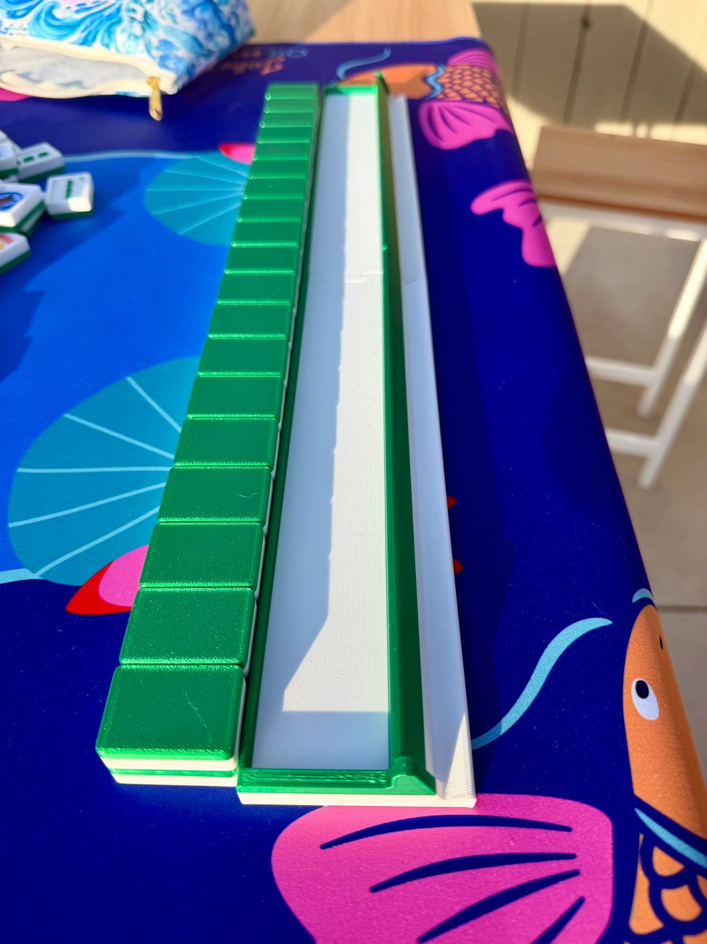 An empty green mahjong rack sits alone on a koi-print mat, its groove ready to hold Travel Mahjong Tiles - 3D.
