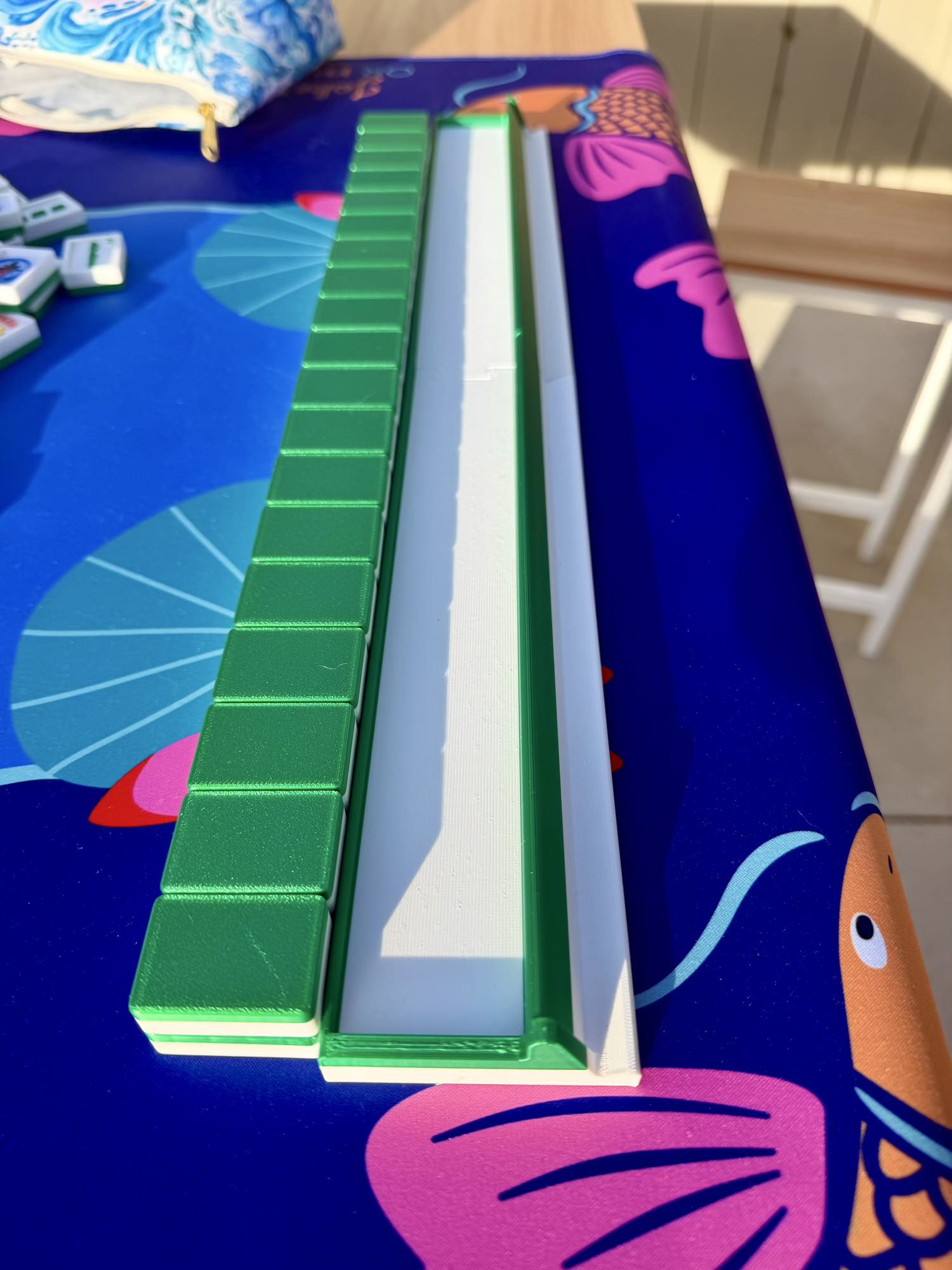 An empty green mahjong rack sits alone on a koi-print mat, its groove ready to hold Travel Mahjong Tiles - 3D.