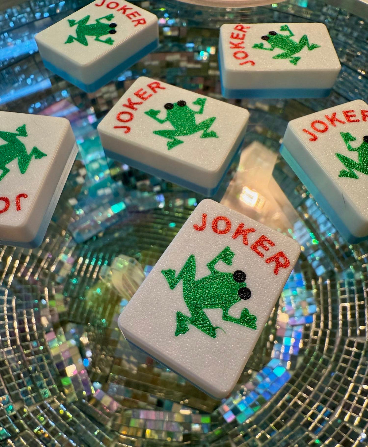 Custom Jokers - Frogs tiles rest on a mirrored disco-ball surface, their green frogs multiplied in the reflection.
