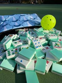 A yellow pickleball rests atop a mint-green pile of Travel Mahjong Tiles spilling from a scalloped pouch on a tennis court.
