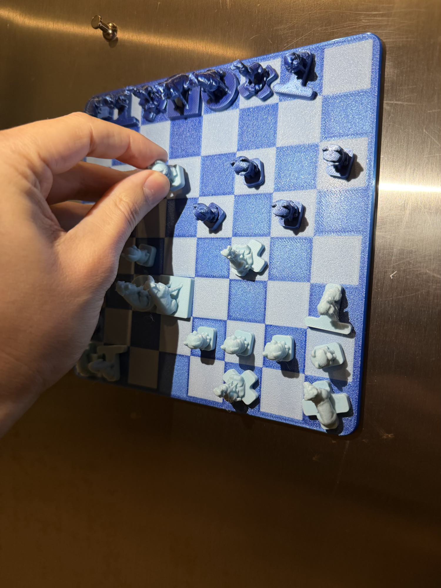 The magnetic Fridge Chess board sticks flat to a stainless steel fridge as a hand moves one pale blue piece into play.
