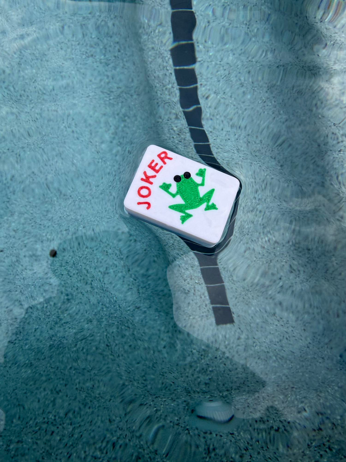 Single frog Joker mahjong tile floating in a pool beside a dark tile lane line, viewed from above