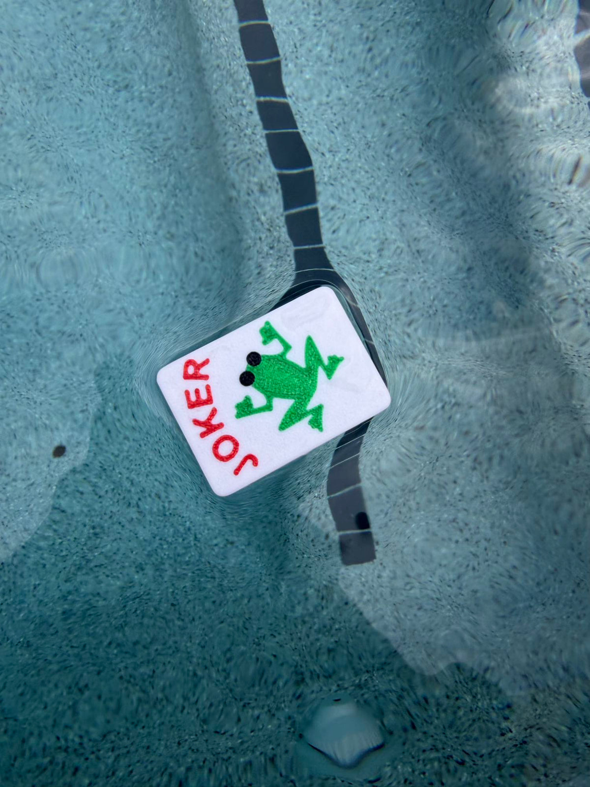 Frog Joker mahjong tile floating upside down in the pool next to the same dark tile lane line