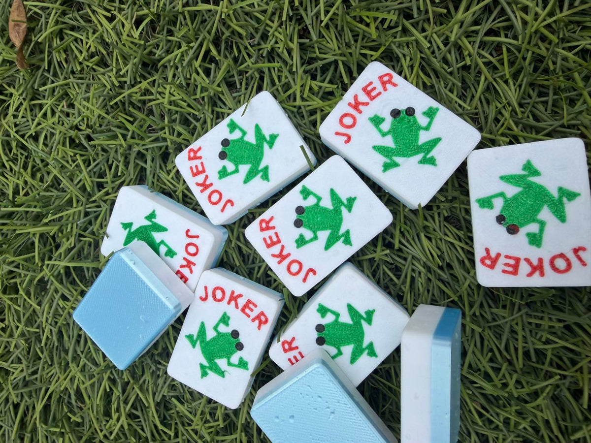 Eight frog Joker mahjong tiles scattered across artificial turf, some paired with plain blue tiles